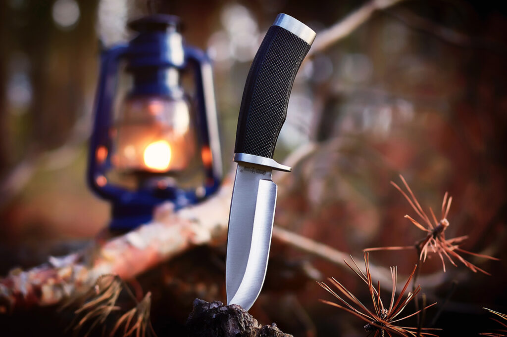 Ein feststehendes Survival-Messer mit schwarzen Griff steckt in einem Baum, im Hintergrund steht eine blaue Sturmlaterne