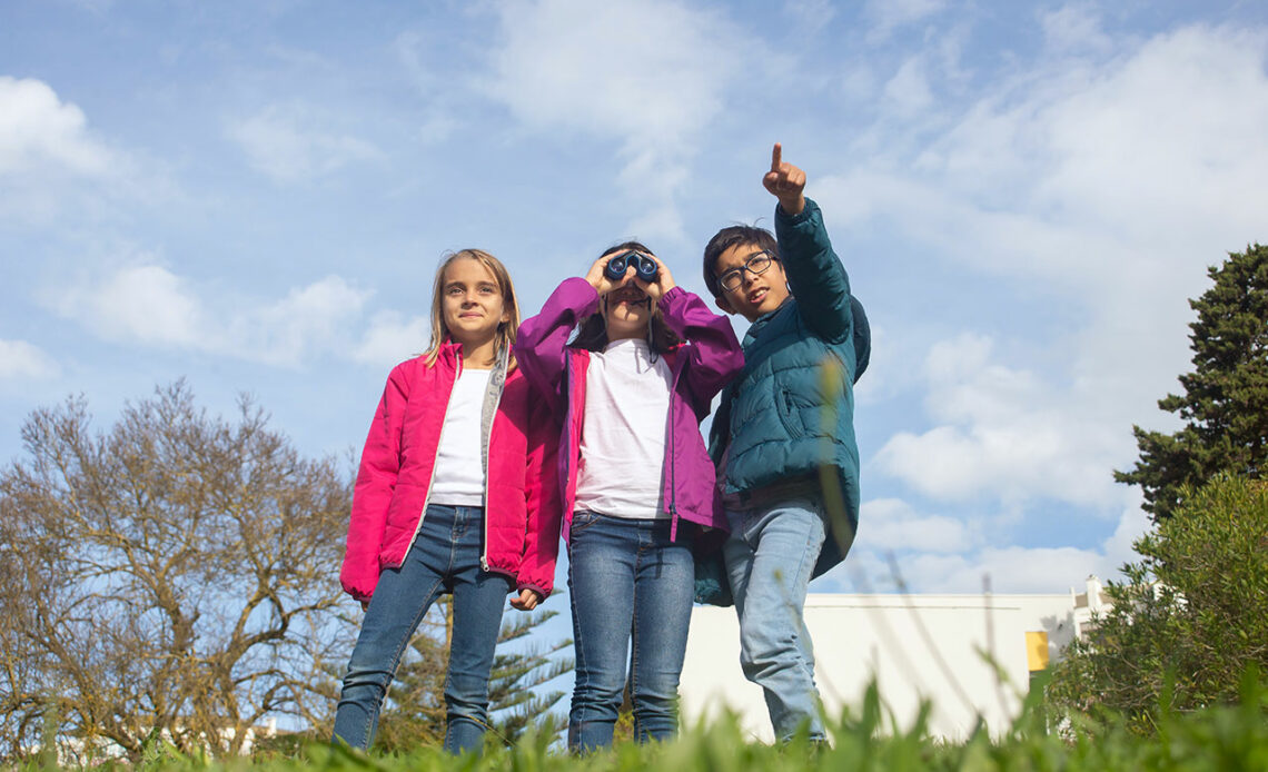 Drei Kinder stehen auf einer Wiese und beobachten die Natur durch ein Fernglas