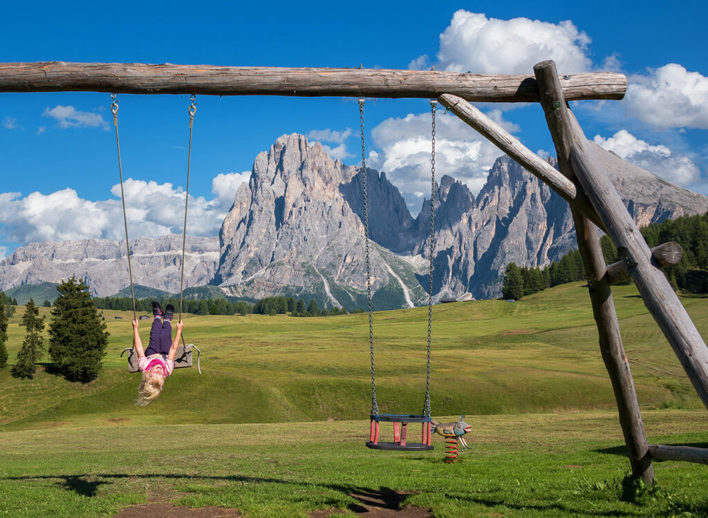 Ein Kind schaukelt auf einem Spielplatz, im Hintergrund ein Bergpanorama