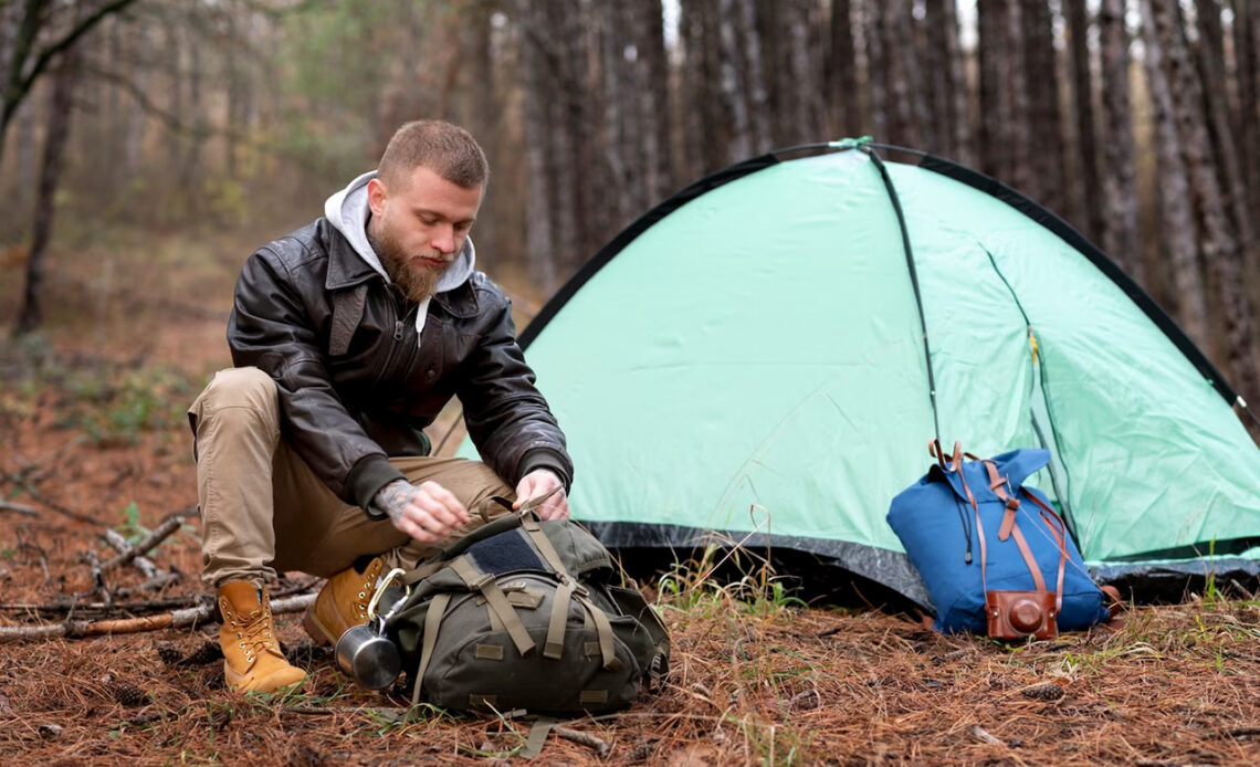 Ein junger Mann hat sein hellblaues Zelt im Wald aufgebaut und packt seinen Wanderrucksack