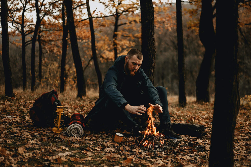 Ein Mann hat sich für die Nacht ein Biwak eingerichtet und wärmt die Hände an einer kleinen Feuerstelle