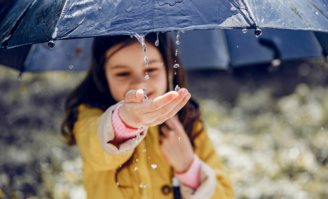 Ein Mädchen mit einer gelben Jacke fängt mit der rechten Hand den Regen vom Rand ihres Regenschirms auf